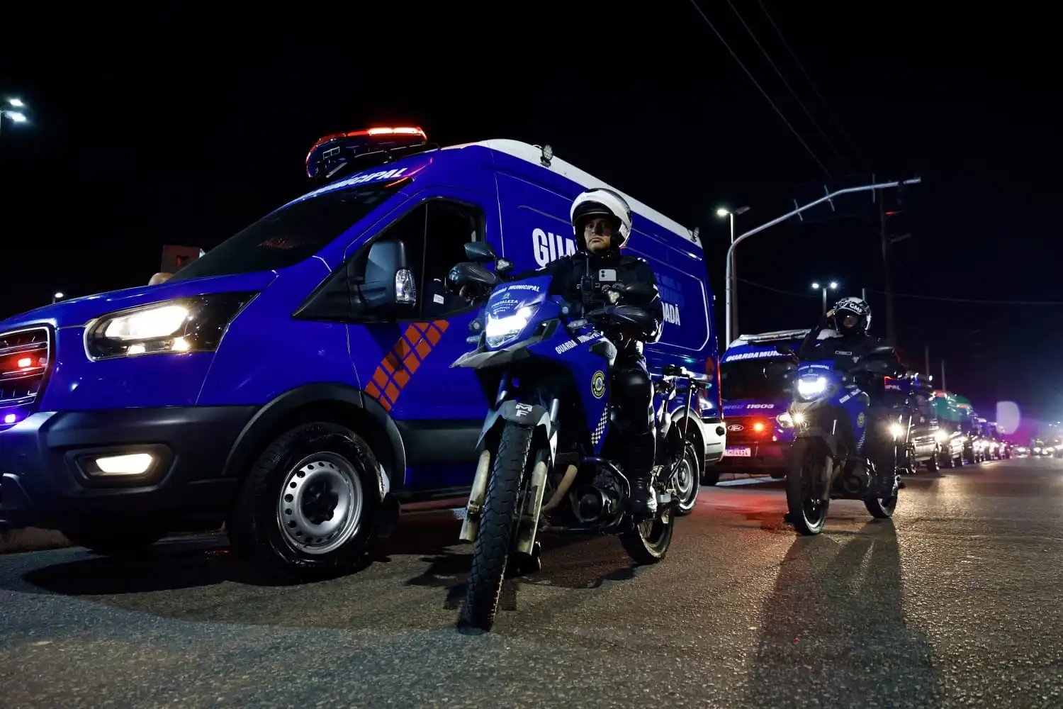 A foto mostra uma van dos Pelotões dos Bairros trafegando por uma rua junto a guardas pilotando motocicletas do programa