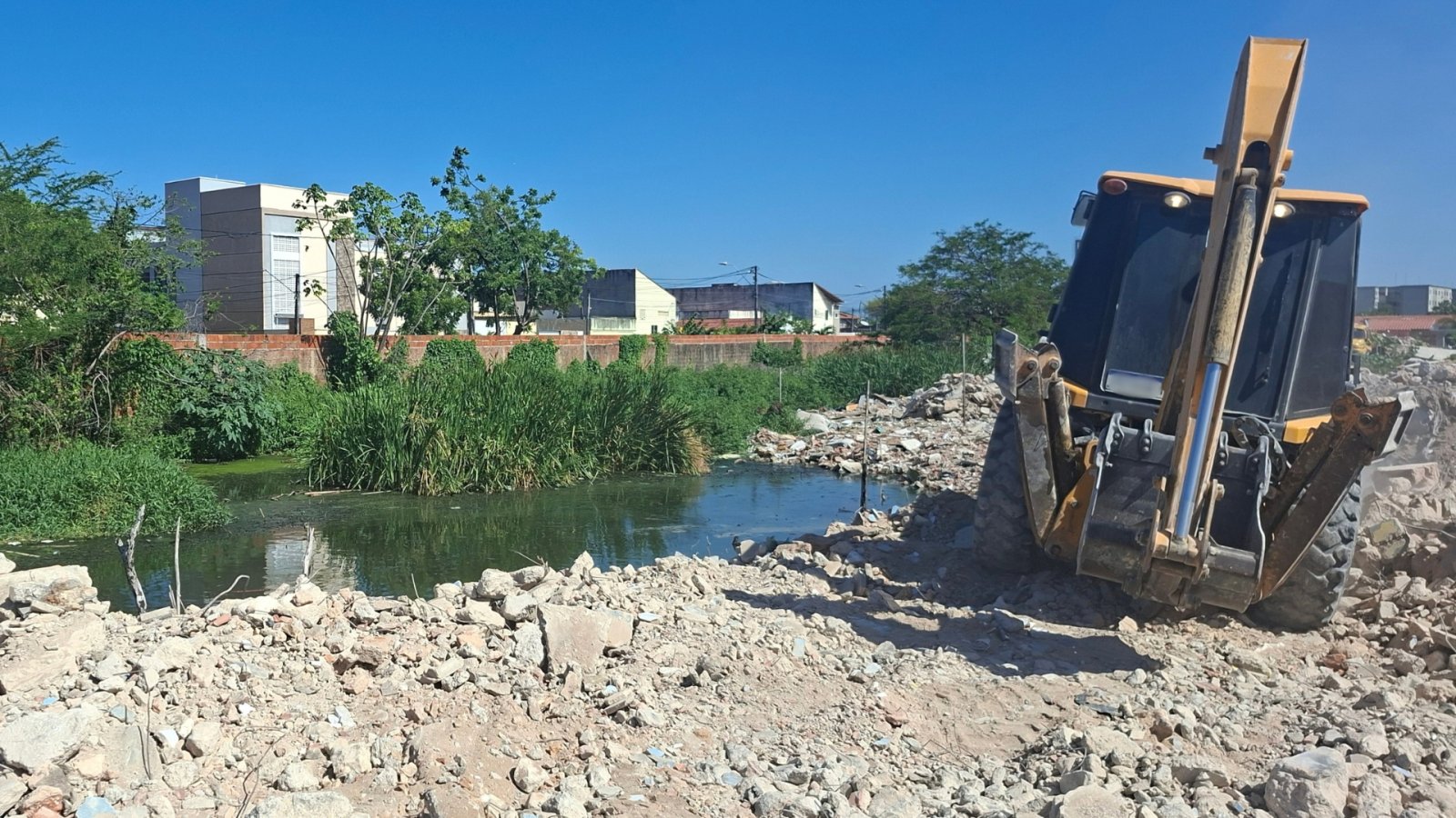 Imagem mostra retroescavadeira à frente de entulho de obras e às margens de rio